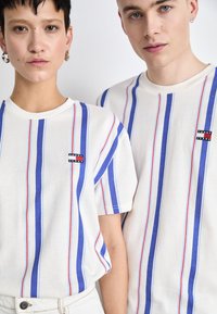 T-shirts en coton blanc avec des rayures verticales bleues et rouges. Chaque T-shirt arbore un petit logo noir "TOMMY JEANS" sur la poitrine.