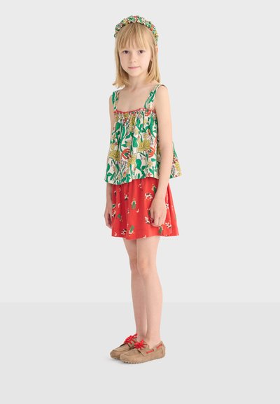 Jeune fille debout portant un haut sans manches vert à fleurs blanches, une jupe rouge avec des imprimés d'animaux, un serre-tête assorti, et des mocassins marron avec des lacets rouges.
