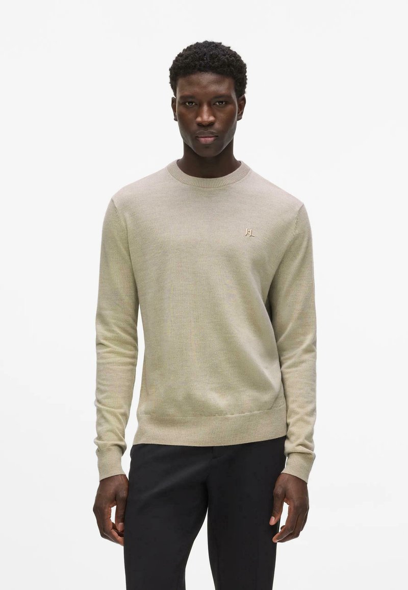 Jeune homme debout, portant un pull beige à manches longues avec un petit logo sur la poitrine et un pantalon noir, expression neutre, fond blanc uni.