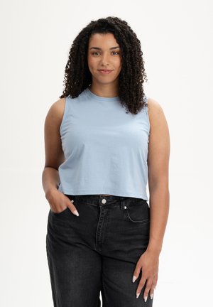 Jeune femme aux cheveux bouclés portant un haut sans manches bleu clair et un jean noir, debout avec une main dans la poche, fond neutre.