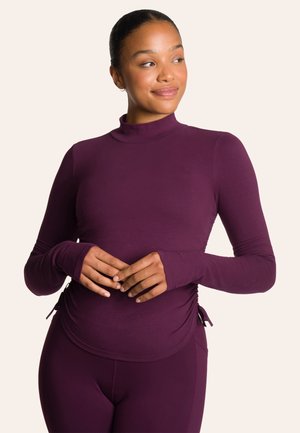 Haut à manches longues et col montant en tissu violet profond. Présente un design ajusté avec un gathering sur les côtés et des trous pour les pouces au niveau des poignets. Texture lisse.