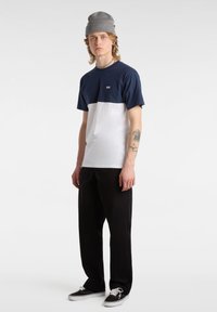 Giovane uomo che indossa un berretto grigio, una maglietta Vans blu e bianca, pantaloni neri e scarpe da ginnastica nere, in piedi con espressione neutra su sfondo bianco.