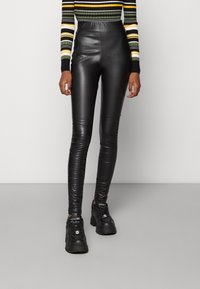 Person som bär svarta höga leggings i konstläder, svarta klumpiga sneakers och en randig långärmad topp med gula, vita och gröna band.