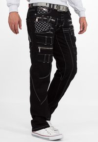 Pantaloni cargo neri con cuciture bianche a contrasto, molteplici tasche con zip e dettagli testurizzati, abbinati a sneakers bianche.