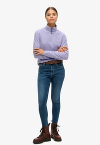 Licht paarse fleece pullover met een halve rits kraag, gedragen over blauwe skinny jeans en bruine veterschoenen. Eenvoudige logo-accenten.