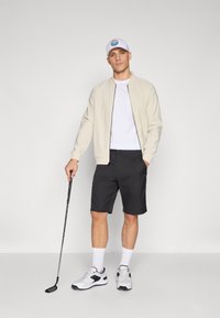 Beige bomber jacket over a white t-shirt, paired with black shorts. White cap and athletic shoes with black accents, holding a golf club.