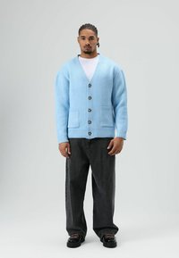 Cardigan bleu clair en tricot avec un col en V, six boutons et deux poches avant, associé à un jean ample foncé et des chaussures noires.