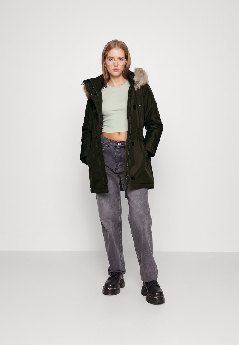 Parka verde oliva con finiture in pelliccia sintetica, tasche frontali, bottoni e zip, abbinata a un crop top verde chiaro e pantaloni grigi.