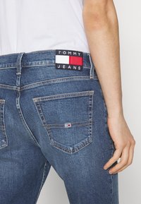 Jeans de denim azul con un corte clásico. Presenta una etiqueta negra con acentos en rojo, blanco y azul en la parte trasera, además de un diseño de bolsillo estándar.
