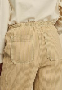 Pantaloni bej în dungi cu talie ajustabilă, având două buzunare pe spate și un material texturat. Dungile sunt de culoare verde deschis și bej.