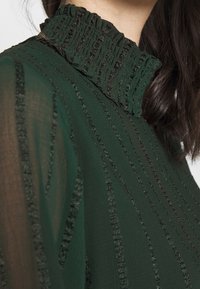 Blouse verte texturée avec un col haut volanté et des motifs à rayures verticales d'un éclat subtil, mettant en valeur un design de tissu superposé.