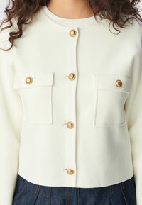 Cardigan blanc à boutons avec deux poches poitrine, présentant des boutons dorés, porté avec un pantalon plissé bleu foncé.