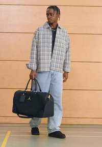 Sort duffelbag med gullfarget metallbeslag, rutete oversized skjorte i grått og hvitt, svart t-skjorte, lyseblå jeans og svarte sko. Glatte teksturer.