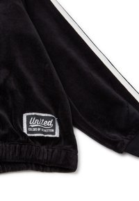 Giacca in velluto nero con strisce bianche ai lati, polsini elastici e etichetta "United Colors of Benetton" su una toppa argentata sulla manica.