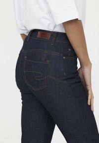 Donkerblauwe spijkerbroek van denim met oranje stiksels, voorzien van een leren logo-patch op de tailleband en een ontwerp op de achterzak. Aansluitende stijl.