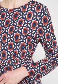 Robe bleu marine à motifs floraux en rouge, blanc et bleu. Le tissu est lisse avec des manches longues et un col rond.