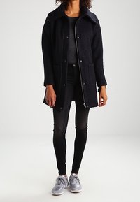 Abrigo de lana azul marino oscuro con un gran cuello, botones a presión y dos bolsillos frontales, llevado sobre una camiseta gris y jeans negros, combinado con zapatillas grises.
