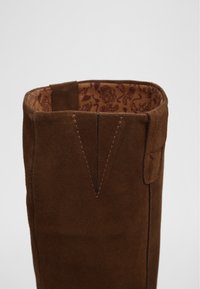Bruine suede laars met een open bovenkant, voorzien van een stikseldetail en een binnenste bloemenpatroon. Glad textuur en gestructureerde vorm met zijtab.