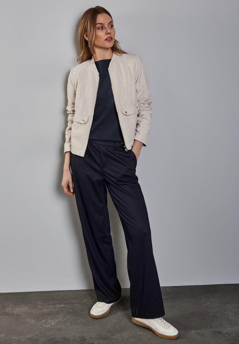 Beige leichte Jacke mit zwei Fronttaschen, kombiniert mit navyblauen, gestreiften, weiten Hosen und weißen Sneakers auf einem grauen Hintergrund.