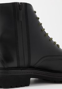 Bottillon en cuir noir avec une fermeture éclair latérale, des lacets jaunes et noirs, et une semelle texturée ; présente une finition lisse et des détails de couture.