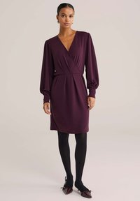 Donna che indossa un abito avvolgente a maniche lunghe color prugna con collant neri e ballerine a punta prugna, in piedi davanti a uno sfondo beige semplice.