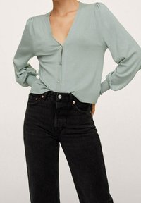 Miękka zielona bluzka z długim rękawem, dekoltem w serek, detalami z guzikami i bufkami na ramionach, w połączeniu z czarnymi, wysokim stanem jeansami.