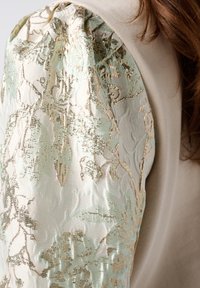 Haut à manches longues avec un corps en crème lisse et des manches bouffantes à motifs floraux texturés en vert pâle et or, avec un tissu froncé.