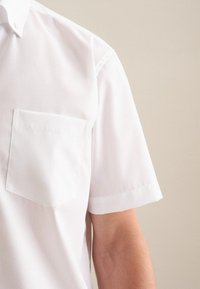 Chemise blanche à manches courtes en tissu lisse, dotée d'une poche poitrine unique et d'un col boutonné avec des coutures nettes et structurées.