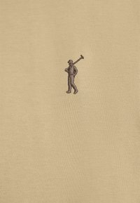 Beżowa koszula z krótkim rękawem, ozdobiona haftowanym brązowym motywem trzymającym narzędzie, umieszczonym po lewej stronie klatki piersiowej. Gładka tekstura.