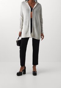 Lehký kardigan s otevřeným pletením v krémové barvě, s kapucí, dlouhými rukávy a uvolněným střihem. Kombinovaný s černými upnutými kalhotami a platformovými botami.