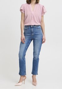 Jasnoróżowa bluzka w kropki z krótkimi falbanianymi rękawami, zestawiona z prostymi dżinsami o średnim odcieniu i bladymi różowymi szpiczastymi szpilkami.