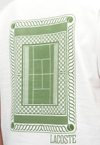 Λευκό μπλουζάκι με εκτύπωση ενός πράσινου γηπέδου τένις, περιγραμμένο με διακοσμητικά σχοινιά και πλέγματα, και το όνομα της μάρκας "LACOSTE" από κάτω.