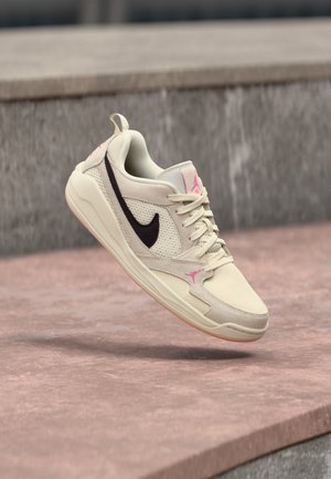 Beiger Nike-Sneaker mit schwarzem Swoosh und pinken Akzenten, schwebend über einer rosa-kräftigen Betonoberfläche vor einem Hintergrund aus grauer Steinmauer.