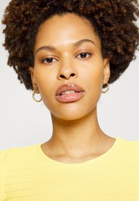 Top amarelo texturizado com mangas curtas em puff, usado por uma pessoa com cabelo encaracolado. Brincos de argola dourados e cor de lábios brilhante são visíveis.