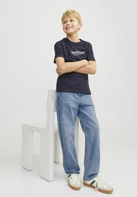 Camiseta de algodón navy con logo blanco, combinada con jeans de mezclilla azul claro. El modelo lleva zapatillas blancas con detalles verdes, de pie de manera casual.