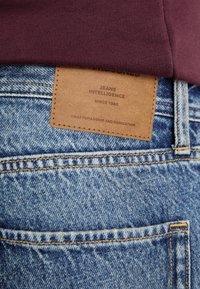 D�žíny z denimu s modrým umytím, s koženou etiketou přišitou k pasu, na které je napsáno "Jeans Intelligence" a informace o řemeslném zpracování.