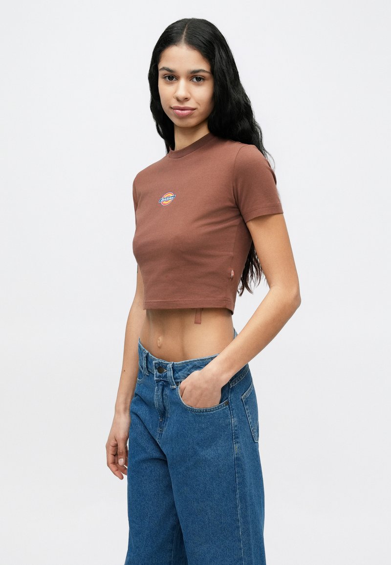 Vrouw met lang donker haar, gekleed in een bruin cropped T-shirt en blauwe spijkerbroek, met één hand in de zak, staand tegen een effen witte achtergrond.