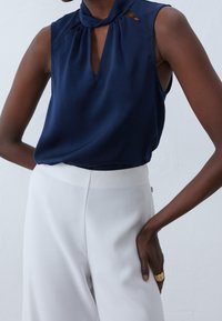 Granatowa bezrękawnikowa bluzka z kołnierzykiem, kluczykowym wycięciem z przodu i plisowanymi akcentami. Połączona z białymi spodniami o wysokim stanie. Gładka tekstura materiału.
