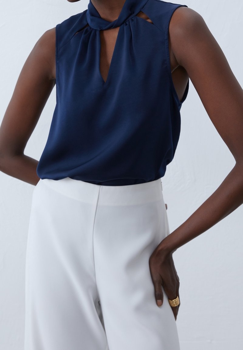 Granatowa bezrękawnikowa bluzka z kołnierzykiem, kluczykowym wycięciem z przodu i plisowanymi akcentami. Połączona z białymi spodniami o wysokim stanie. Gładka tekstura materiału.