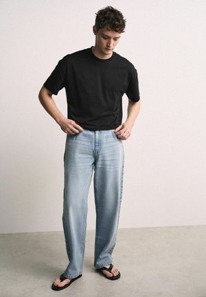 Junger Mann trägt ein schwarzes T-Shirt, hellblaue Jeans mit weitem Bein und schwarze Flip-Flops, steht und passt die Bundweite seiner Hose an.