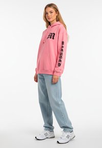 Felpa rosa con testo grafico nero, tasca frontale e cappuccio con cordino, abbinata a jeans baggy azzurri e scarpe sportive bianche.