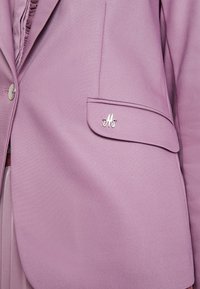 Lniany blezer w kolorze bzu, wykonany z gładkiej tkaniny, z pojedynczym zapięciem na guziki oraz kieszenią z klapą ozdobioną srebrnym logo "M".