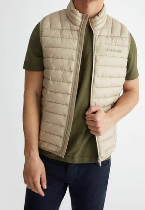 Man draagt een beige doorgestikt vest over een groen kortärmelig shirt, houdt met één hand de rits van het vest vast, tegen een effen witte achtergrond.