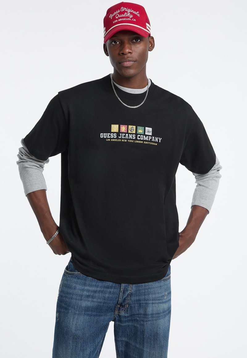 Maglietta nera a maniche corte con maniche lunghe grigie sotto, presenta un logo multicolore e il testo "GUESS JEANS COMPANY". Abbinata a jeans blu.