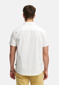 Camisa blanca de manga corta con tejido texturizado, cuello puntiagudo y ajuste holgado, combinada con pantalones amarillo claro, vista desde atrás.