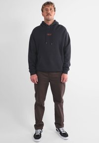 Sweat à capuche noir avec cordons de serrage et poche avant, associé à un pantalon marron et des baskets noires. Le sweat à capuche présente un petit logo.