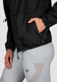 Sort letvægtsjakke med justerbar bund og manchetter, udstyret med en frontlomme; parret med lysegrå leggings med geometriske accenter.