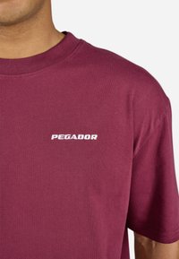 Burgunderfarbener Baumwoll-T-Shirt mit kurzen Ärmeln und Rundhalsausschnitt. Mit weißem, gesticktem "PEGADOR"-Logo auf der Brust. Weicher Stoff.
