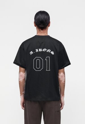 Osoba s vitiligom nosí čierne tričko A. IRONS 01, stojí chrbtom k divákovi, vlasy má zviazané v nízkom drdole, na jednoliatom pozadí.