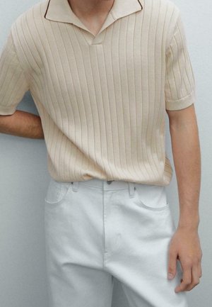 Mann trägt beige, kurzärmeliges, geripptes Strick-Poloshirt, in hoch taillierte weiße Hose gesteckt, steht vor einer schlichten hellen Wand.